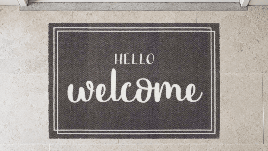 Graue Fußmatte mit weißem „HELLO welcome“-Motiv auf Steinboden